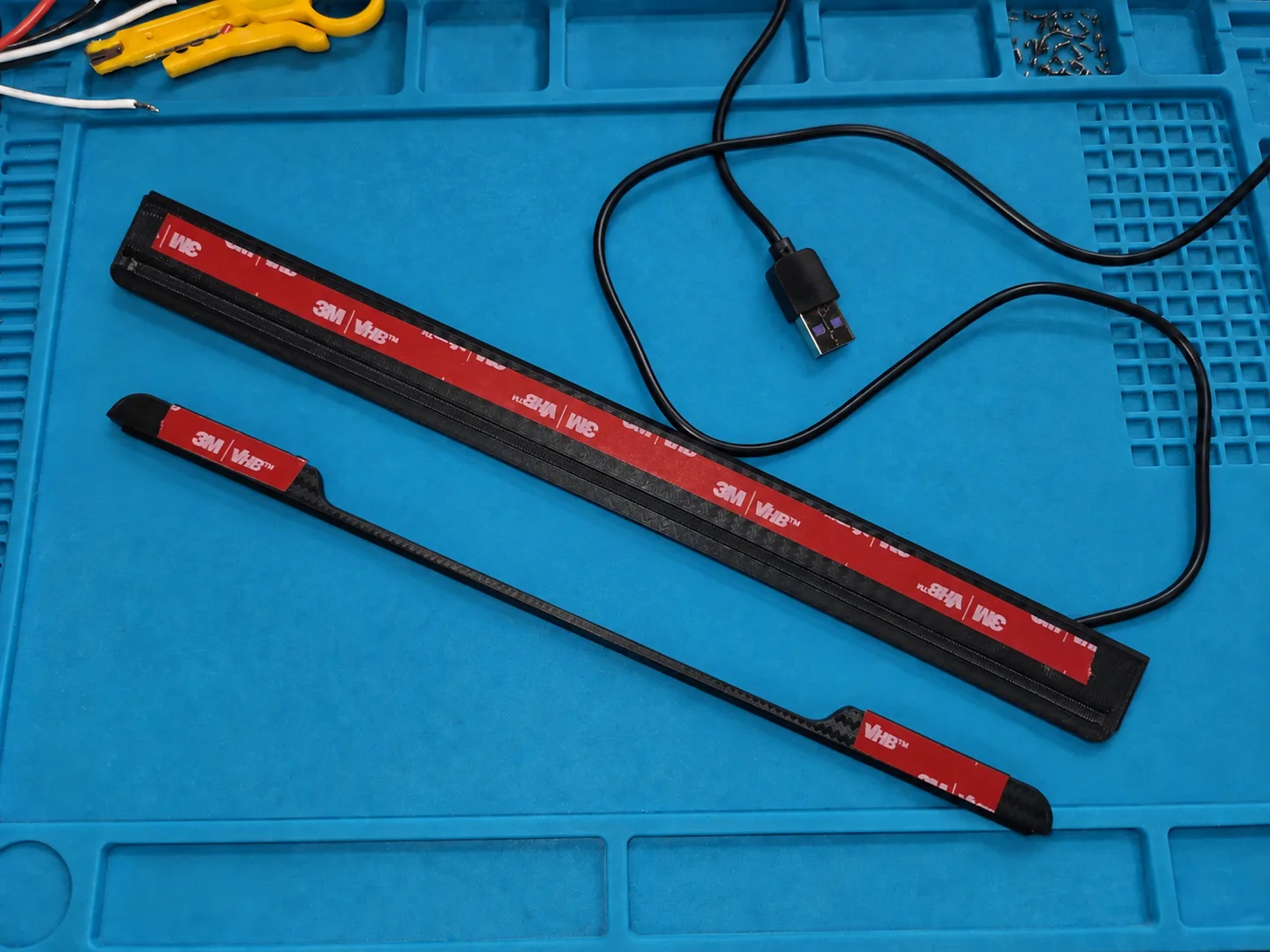 Zwei schwarze Halteteile mit rotem 3M-VHB-Klebeband, daneben ein USB-Kabel.